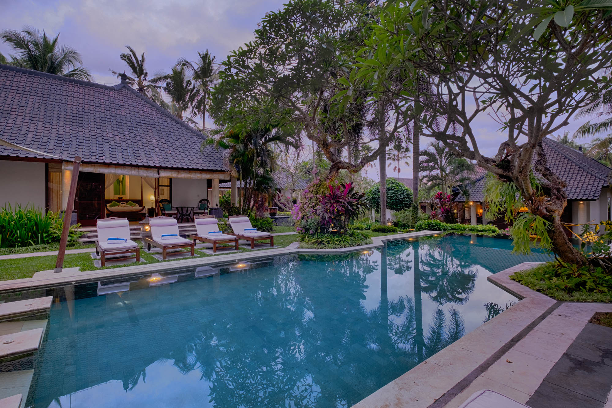 Garden pool with sun loungers beside a tiled-roof villa amid lush tropical planting at twilight, Sanga Sanga, Bali