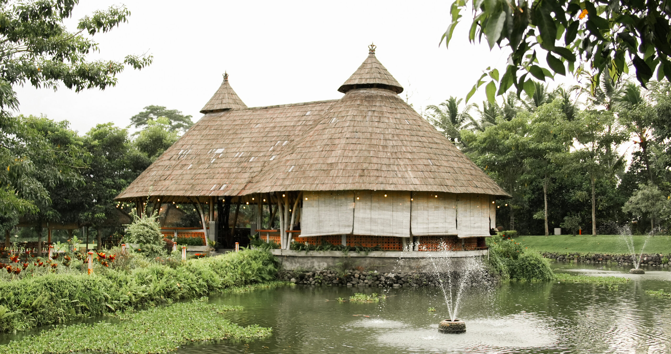 Twin-peaked thatched bamboo restaurant pavilion beside a lake with fountain, Mango Lango Lake Resto